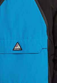 Cazadora de cuello redondo en azul y negro con un parche de logo triangular y un bolsillo frontal con cremallera, fabricada en un tejido suave y ligero.