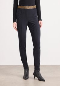 Pantalons noirs fuselés avec une ceinture en imprimé léopard, présentant des coutures de poches avant et une texture lisse, associés à des bottes noires à la cheville.