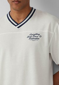 Witte katoenen t-shirt met een v-hals en een marineblauw gestreepte kraag. Geprinte tekst "Alles draait om motivatie" in marineblauw. Getextureerde stof.