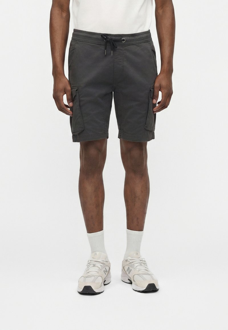 Hombre vistiendo pantalones cortos cargo gris oscuro, calcetines blancos y zapatillas deportivas blancas, de pie contra un fondo blanco liso.