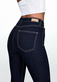 Vaqueros de mezclilla azul oscuro con corte ajustado, que presentan una cintura alta, costuras amarillas y una etiqueta de cuero con la marca en la parte posterior de la cintura.