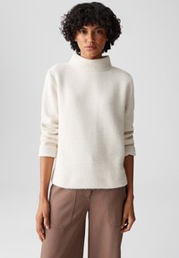 Maglione color crema con collo alto, tessuto strutturato e maniche a tre quarti, abbinato a pantaloni a gamba larga marroni.