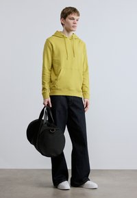 Κίτρινο φούτερ με κουκούλα και μπροστινή τσέπη, συνδυασμένο με σκούρο φαρδύ τζιν. Μαύρη στρογγυλή τσάντα duffel και λευκά αθλητικά παπούτσια ολοκληρώνουν το σύνολο.