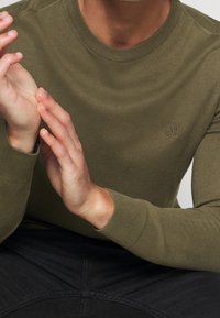 Pull en tricot vert olive avec un col et des poignets côtelés, mettant en avant un logo subtil sur la poitrine. Gros plan de mains reposant sur les cuisses.