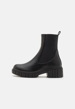 Bottine noire en cuir lisse avec des panneaux latéraux élastiques, des détails texturés et une semelle en caoutchouc épaisse. Boucle au dos.