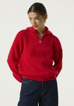 Vrouw met donker haar in een rode trui met ritskraag en donkere jeans staat tegen een effen lichte achtergrond en kijkt licht naar beneden.