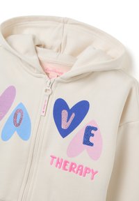 Beige zip-up hoodie met hartvormige ontwerpen in roze, blauw en paarse tinten, met de tekst "LOVE THERAPY" in gestructureerde roze letters.