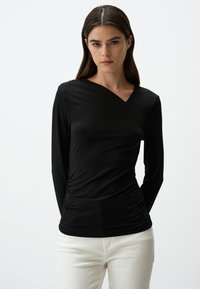 Zwarte long-sleeve top met een crossover v-halsontwerp, gemaakt van gladde stof, met verzamelde textuur bij de taille. Draagt met witte broek.