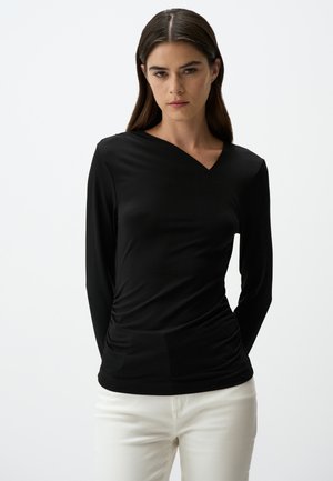Maglia nera a maniche lunghe con design incrociato a V, realizzata in tessuto morbido, con tessuto arricciato in vita. Indossata con pantaloni bianchi.