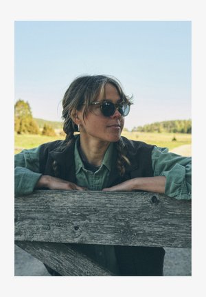 Femeie cu păr castaniu împletit, purtând ochelari de soare rotunzi negri și o cămașă verde cu nasturi, sprijinită de un gard de lemn uzat.