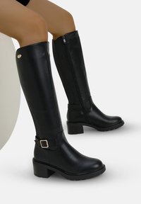 Stivali in pelle nera al ginocchio con tacco a blocco, zip laterale e un accento in metallo sulla fascia. Finitura texturizzata e liscia.