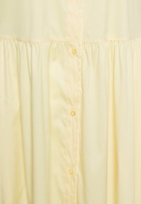 Tissu jaune clair avec fermeture par boutons verticale et détail de couture froncée près du haut.
