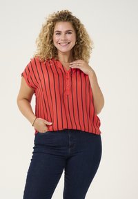 Blusa de rayas rojas con un corte relajado, mangas cortas y cuello de camisa con botones, combinada con unos jeans ajustados de color azul oscuro. Textura de tela suave.