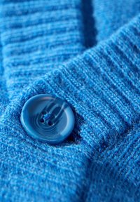 Blauer Strickstoff mit einer strukturierten Musterung und einem großen, glänzenden, runden blauen Knopf mit vier Löchern.