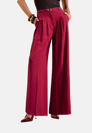 Femme portant un pantalon rouge taille haute à jambes larges avec une petite ceinture, un haut noir, des talons bordeaux et des bracelets, les mains dans les poches.