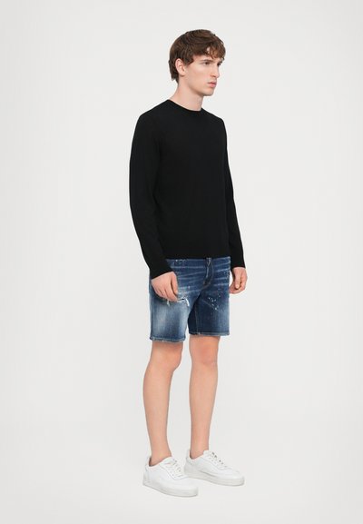 Un pull noir à manches longues, un short en denim avec des détails déchirés et des baskets blanches. La tenue présente une silhouette décontractée et des couleurs contrastées.