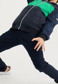 Puffatakki, jossa on vihreä ja laivastonsininen värieste, vetoketjukiinnitys ja ribatut hihansuut; yhdistettynä tummansinihin farkkuihin ja valkoisiin lenkkareihin.