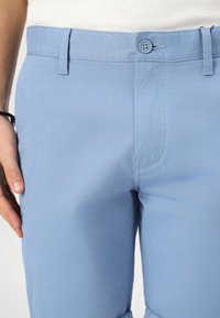 Pantalones azul claro de algodón con una textura suave, con cierre de botón y lazos para el cinturón, mostrando detalles de costura en los bolsillos delanteros.