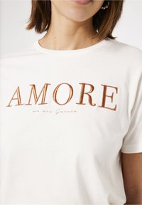 Biała bawełniana koszulka z napisem "AMORE" w brązowym haftem, subtelnym napisem "we are Garcia" poniżej w stylu pisma odręcznego, okrągły dekolt, miękka tekstura.