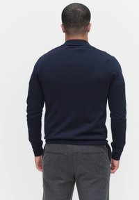 Tummansininen neulepusero, jossa on pitkät hihat ja pyöreä kaula-aukko, yhdistettynä harmaisiin housuihin, joissa on sileä pinta ja istuva muoto.