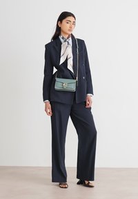 Marineblå, skreddersydd dress med vide bukser, kombinert med et mønstrete silkeskjerf og en liten teal crossover-veske. Sorte åpen-tå sandaler.