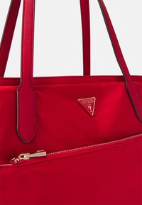 Czerwona torebka wykonana z gładkiego materiału, z złotym trójkątnym logo, dwoma paskami oraz zamykaną na zamek kieszenią z przodu.