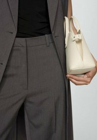 Braune maßgeschneiderte Hose, kombiniert mit einem schwarzen Oberteil, und hält eine kleine cremefarbene Lederhandtasche mit strukturiertem Design und oberen Griffen.