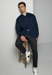 Jachetă bleumarin cu mâneci lungi și nasturi, cu un croi lejer, purtată împreună cu o pereche de pantaloni strâmți de culoare închisă și adidași albi cu accente negre.