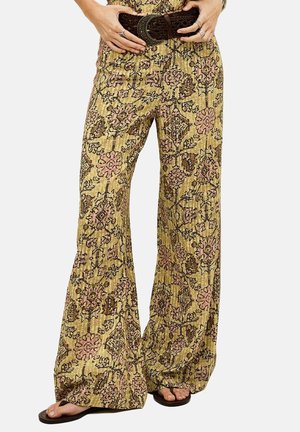 Pantalones amarillos holgados con estampado y diseños florales en rosa y marrón, combinados con un cinturón ancho tejido en marrón y chanclas negras en los pies descalzos.