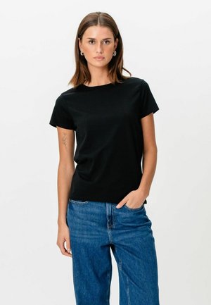 Joven con cabello castaño hasta los hombros, vestida con una camiseta negra de manga corta y jeans azules, de pie con una mano en el bolsillo.
