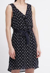 Mörkblå ärmlös klänning med v-ringning, prydd med ett vitt geometriskt triangelmönster och en knytbart midjeband för en definierad silhuett.