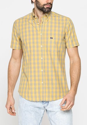 Uomo che indossa una camicia gialla a quadri a maniche corte con taschino sul petto e jeans azzurri, in piedi davanti a uno sfondo semplice.