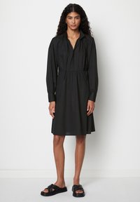 Schwarzes Kleid mit Kragen, langen Ärmeln und geraffter Taille. Hergestellt aus weichem Stoff, hat es eine fließende Silhouette und wird von flachen schwarzen Sandalen ergänzt.