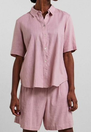 Woman wearing light pink short-sleeve button-up shirt with a collar and matching loose shorts against a plain background.