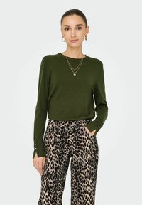 Olivgrüner, langärmeliger Pullover mit dezenten Knopfdetails an den Ärmeln, kombiniert mit weit geschnittenen Leoprint-Hosen mit Taschen.
