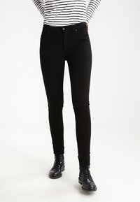 Jeans skinny de mezclilla negra con una textura suave, cintura media, cinco bolsillos y trabillas para cinturón, combinados con botas negras con cordones.