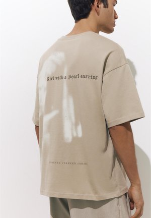 Man draagt een beige oversized t-shirt met de tekst "Meisje met de parel" en "Johannes Vermeer (1632-1675)" op de achterkant.