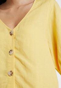 Geel blouse met knopen en een diepe V-hals, gemaakt van lichte stof. Heeft beige knopen met een subtiel patroon aan de voorkant.