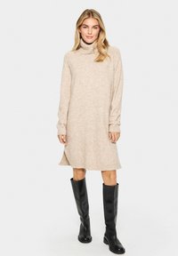 Beige polokrageklänning med långa ärmar, en löst sittande passform och en något utsvängd nederkant; ihop med knähöga svarta stövlar.