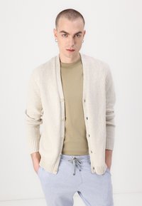 Cardigan tricotat de culoare bej deschis, cu guler tip șal, cinci nasturi și manșete cu nervuri, purtat peste o cămașă de culoare verde măslin și pantaloni gri cu șnur.
