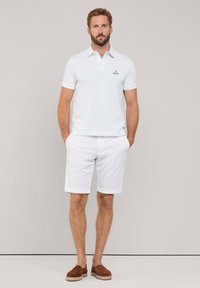 Camisa polo blanca con cuello, logotipo bordado, combinada con pantalones cortos blancos y zapatos marrones sin cordones. Conjunto simple y clásico de verano.