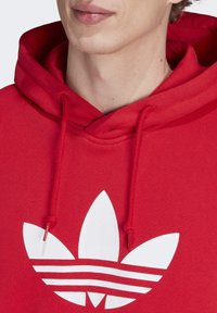 Detailní záběr na osobu v červené mikině s velkým bílým logem Adidas trefoil na hrudi a s tkaničkami volně visícími dolů.