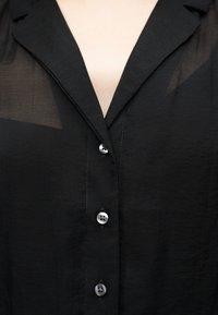 Κοντινό πλάνο ενός ατόμου που φορά μαύρο διάφανο μπλουζάκι με τρία ορατά κουμπιά και βαθύ V-λαιμόκοψη.