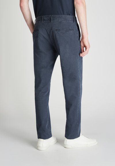 Granatowe spodnie uszyte z gładkiego materiału, oferujące dopasowany krój, dwie tylne kieszenie oraz zamek błyskawiczny. Noszone z białymi trampkami.