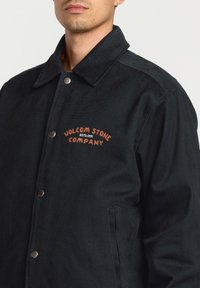 Black canvas jacket with a pointed collar, snap buttons, and an orange embroidered logo on the chest. Features side pockets.