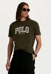 Man som bär en mörkgrön Polo Ralph Lauren t-shirt, svarta cargo-byxor, solglasögon och armbandsur, står med en hand i fickan.