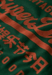 Tessuto verde con lettering e pattern grandi e audaci in arancione. Il testo include sia caratteri inglesi che non inglesi. Texture morbida.