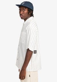 Korte mouwen overhemd in wit met zwarte verticale strepen, voorzien van een borstzak en een zwart logo patch op de mouw.