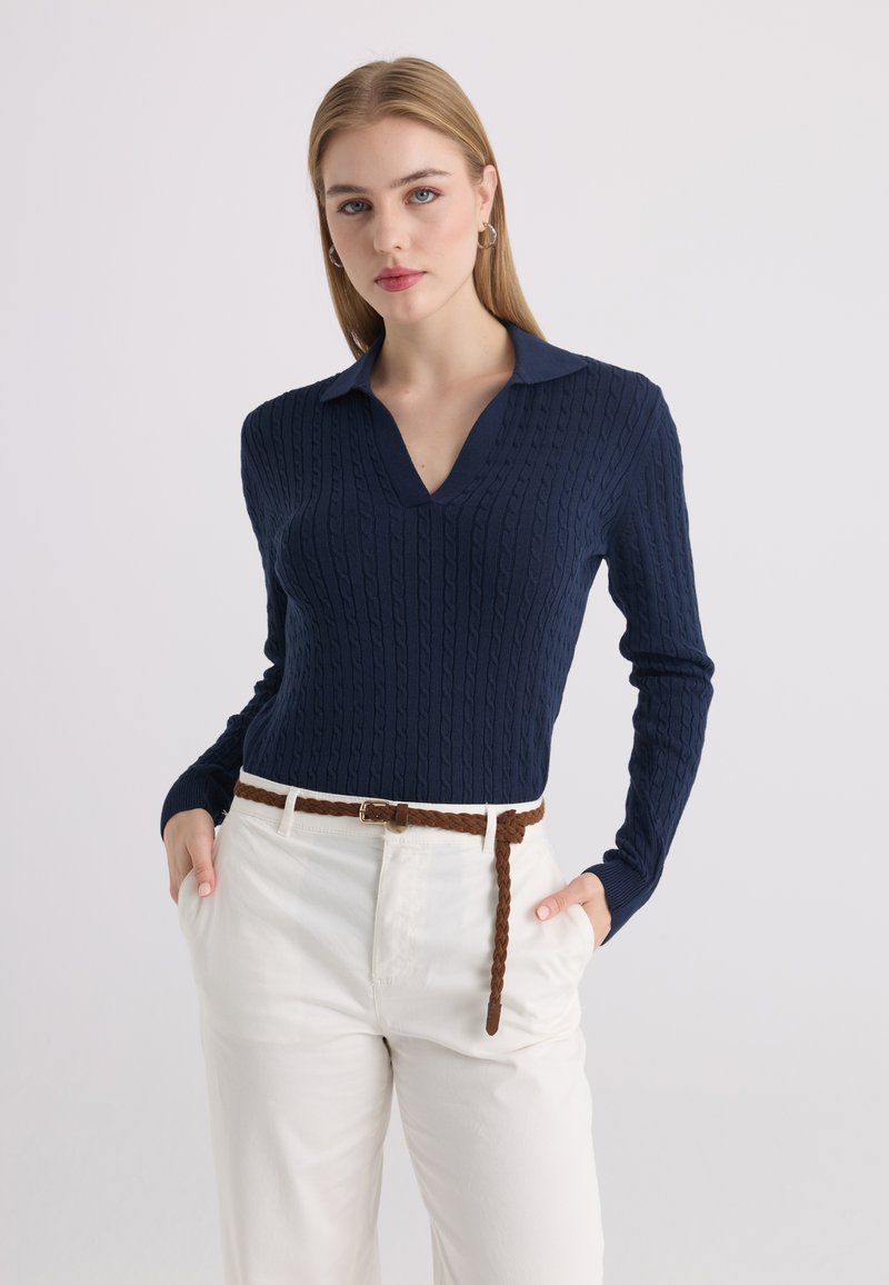 Jeune femme aux cheveux blonds et raides porte un haut bleu marine en maille torsadée à manches longues et un pantalon blanc avec une ceinture tressée marron.