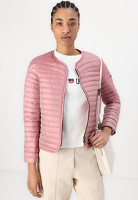 Casaco bomber acolchoado em rosa claro, com decote arredondado, mangas longas e detalhe de fecho preto, usado sobre um sweat branco e calças creme.